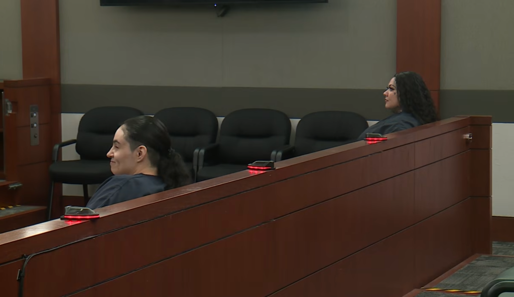 Two women, wearing dark shirts, sitting in a courtroom with wooden railing in front of them.